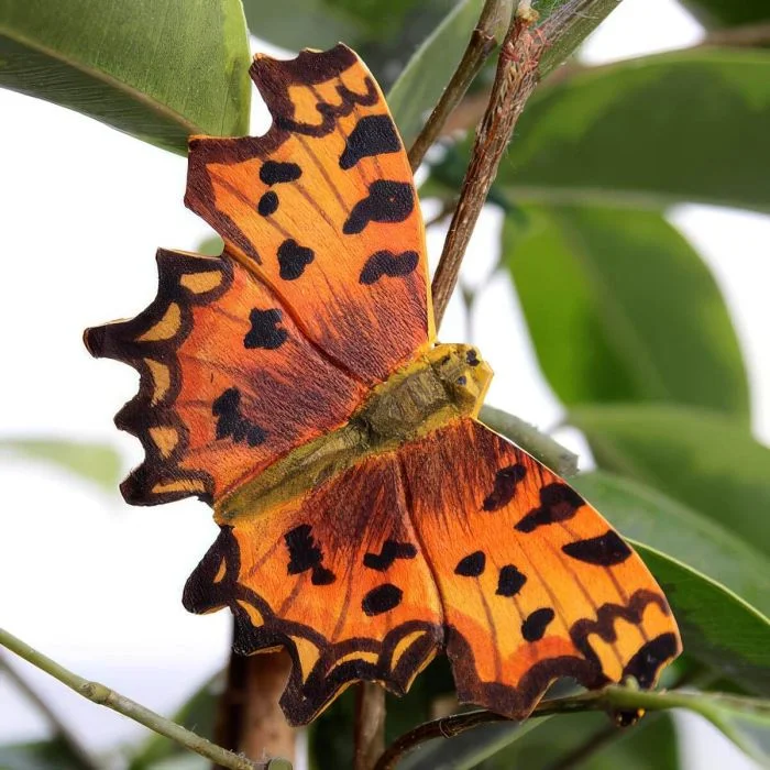 Houten DecoButterfly Gehakkelde aurelia van Wildlife Garden met oranje vleugels, zwarte vlekken en gekartelde vleugelranden, geplaatst tussen groene bladeren.