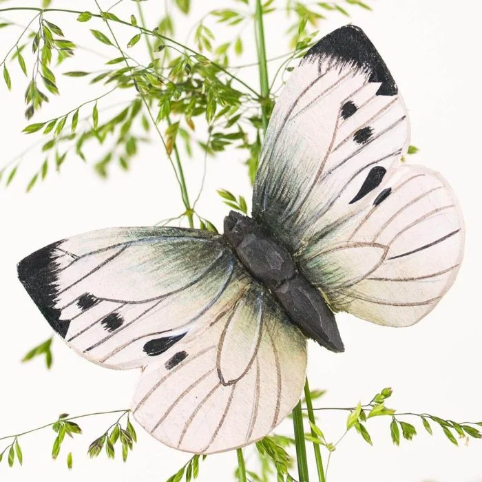 DecoButterfly Groot koolwitje van Wildlife Garden als houten vlinderdecoratie tussen fijne groene takjes, met witte vleugels, zwarte punten en donkere vlekken.