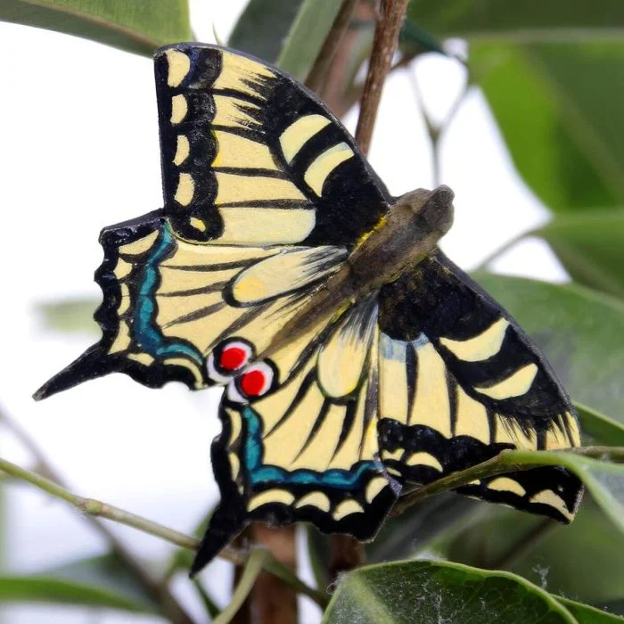 DecoButterfly Koninginnenpage van Wildlife Garden als houten vlinderdecoratie tussen groene bladeren, met geelzwarte vleugels, blauwe accenten en rode oogvlekken.