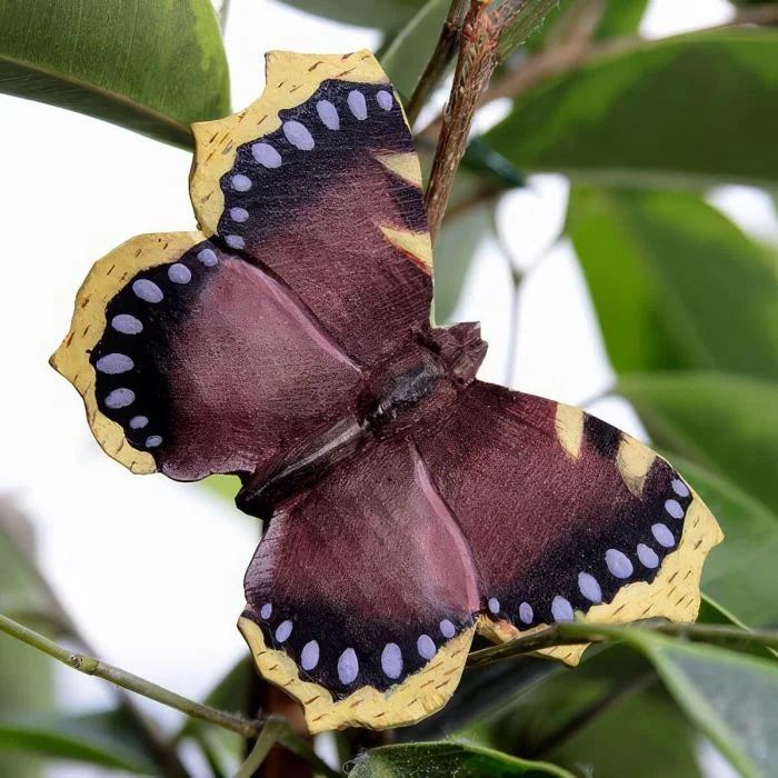 DecoButterfly Rouwmantel van Wildlife Garden als houten vlinderdecoratie tussen groene bladeren, met donkerbruine vleugels, blauwe stippen en gele randen.