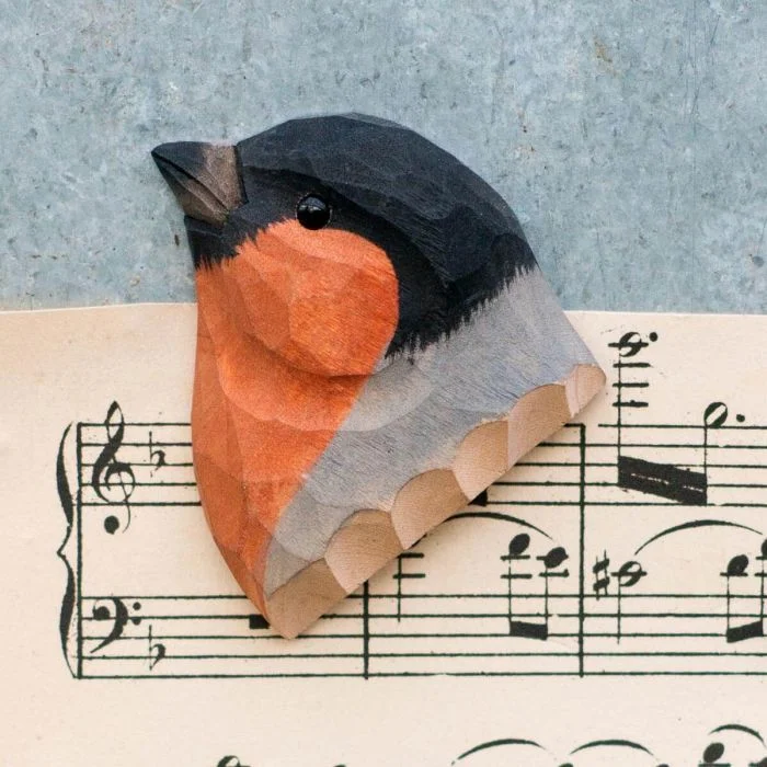 Houten koelkastmagneet Goudvink van Wildlife Garden met zwarte kop, oranjerode borst en grijze vleugels, liggend op een blad met bladmuziek.